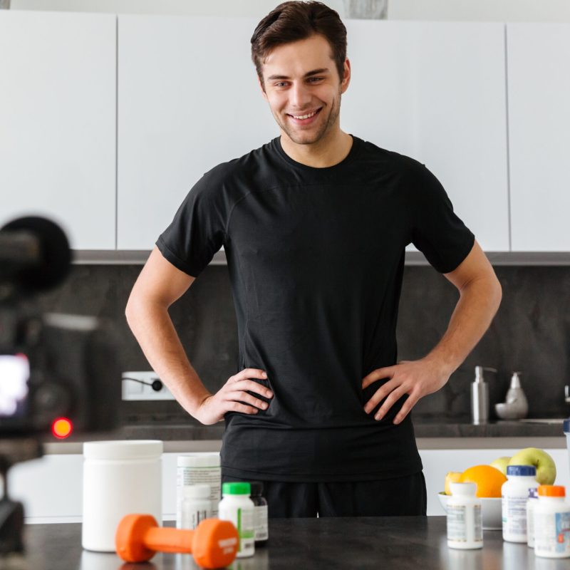 Happy young man filming his video blog episode Happy young man filming his video blog episode about healthy food additives while standing at the kitchen table