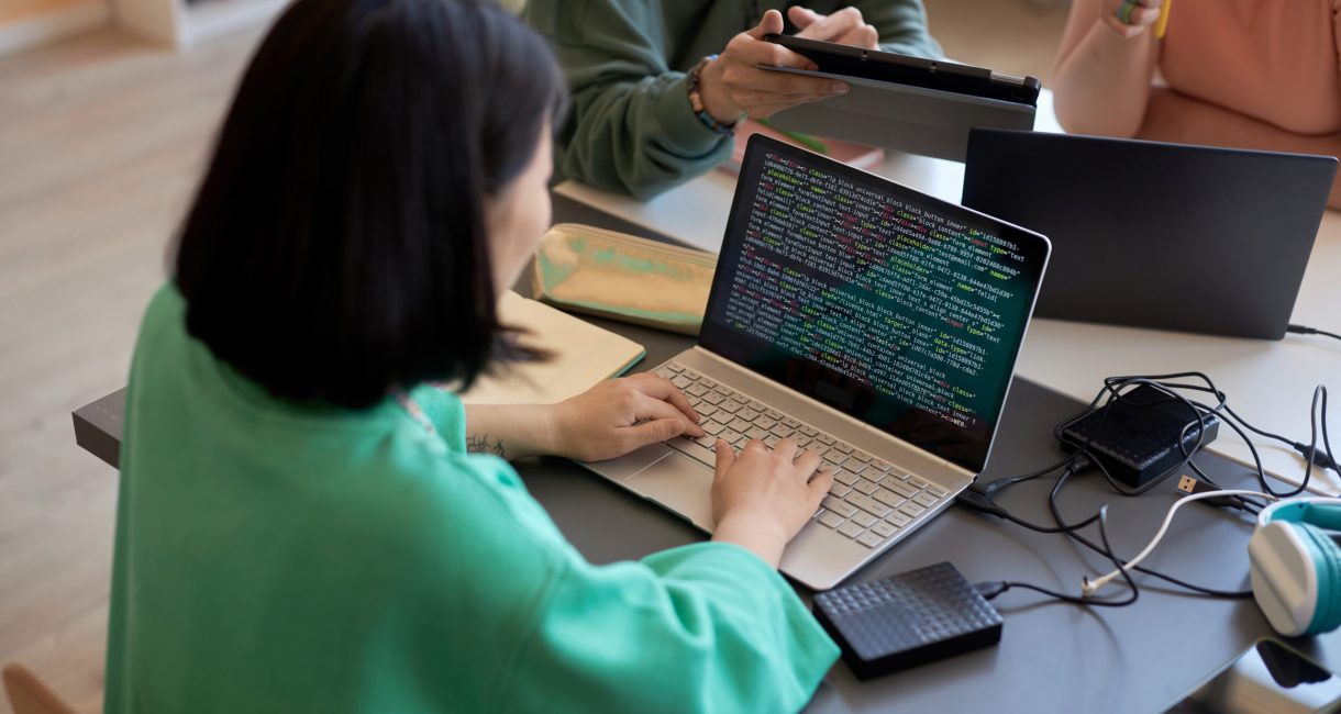 Young female student typing on laptop keyboard while decoding data on screen against two classmates or colleagues discussing presentation